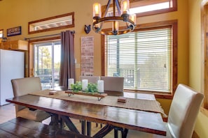 A dining area with a wooden table, cushioned chairs, a chandelier, and a view through large windows.