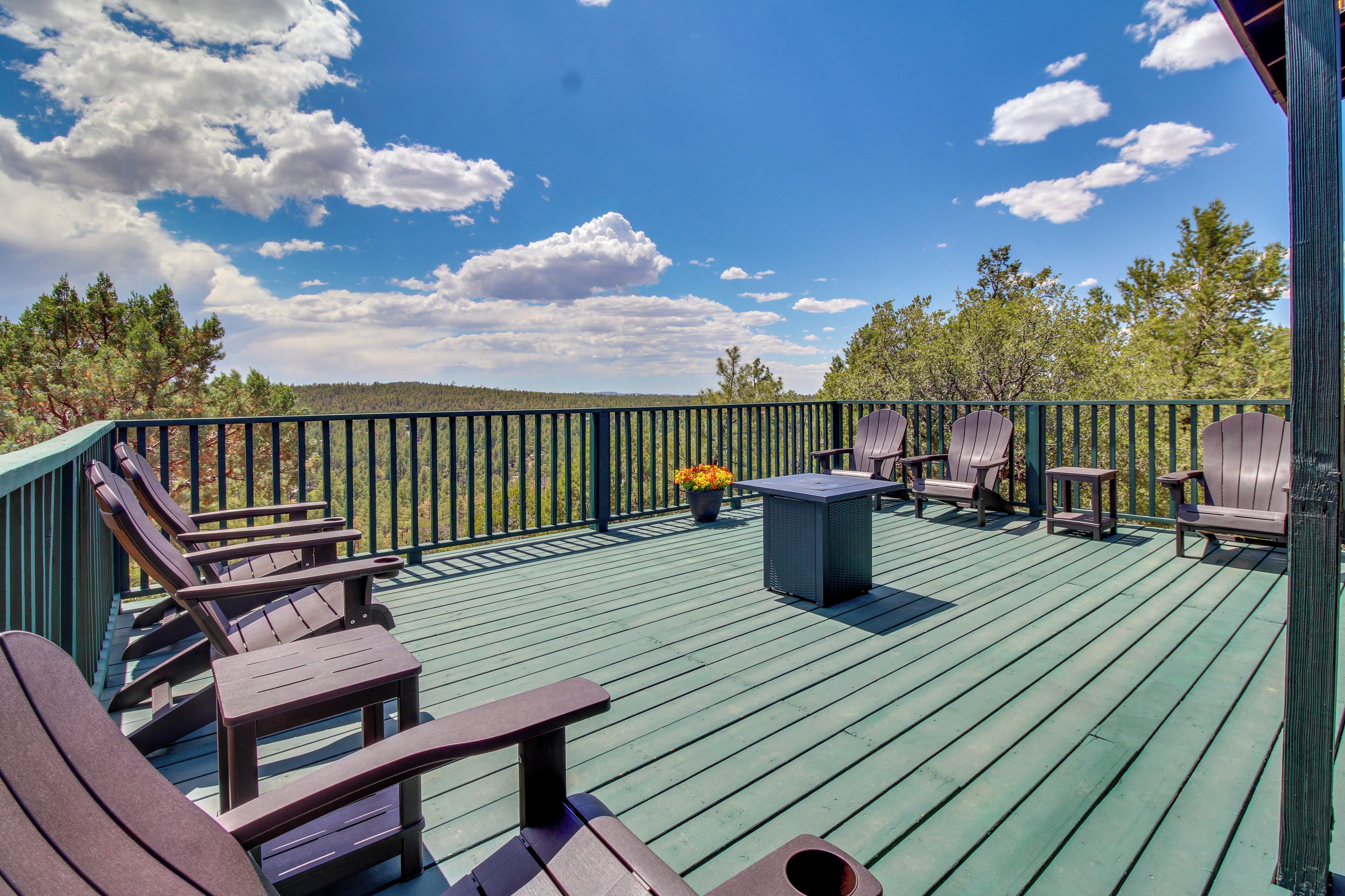 Eine Holzterrasse mit Adirondack-Stühlen und einem Tisch, umgeben von einem Geländer mit Blick auf Bäume und Himmel.