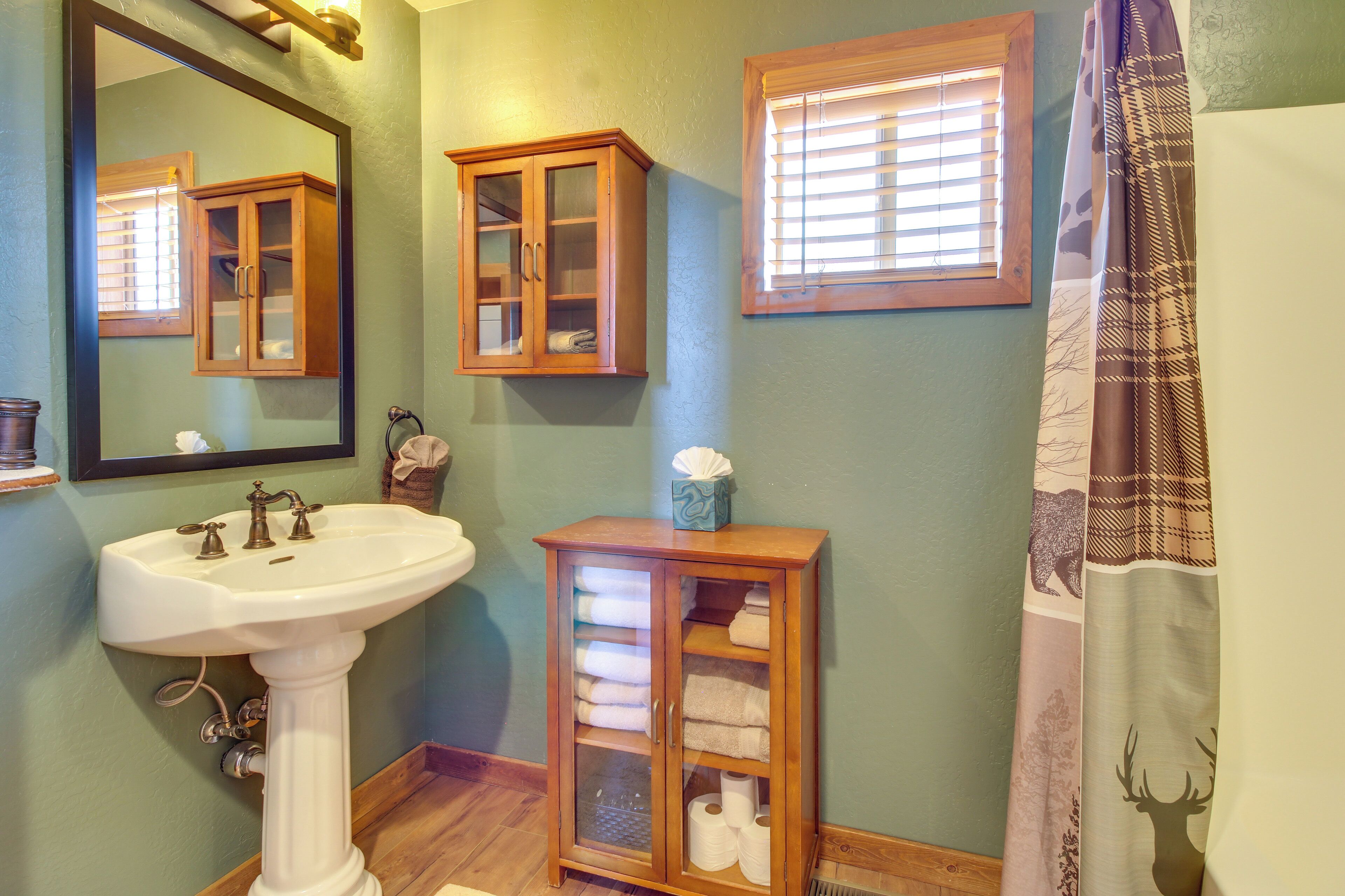 A bathroom with a pedestal sink, two wooden cabinets, a mirror, and a window with blinds.