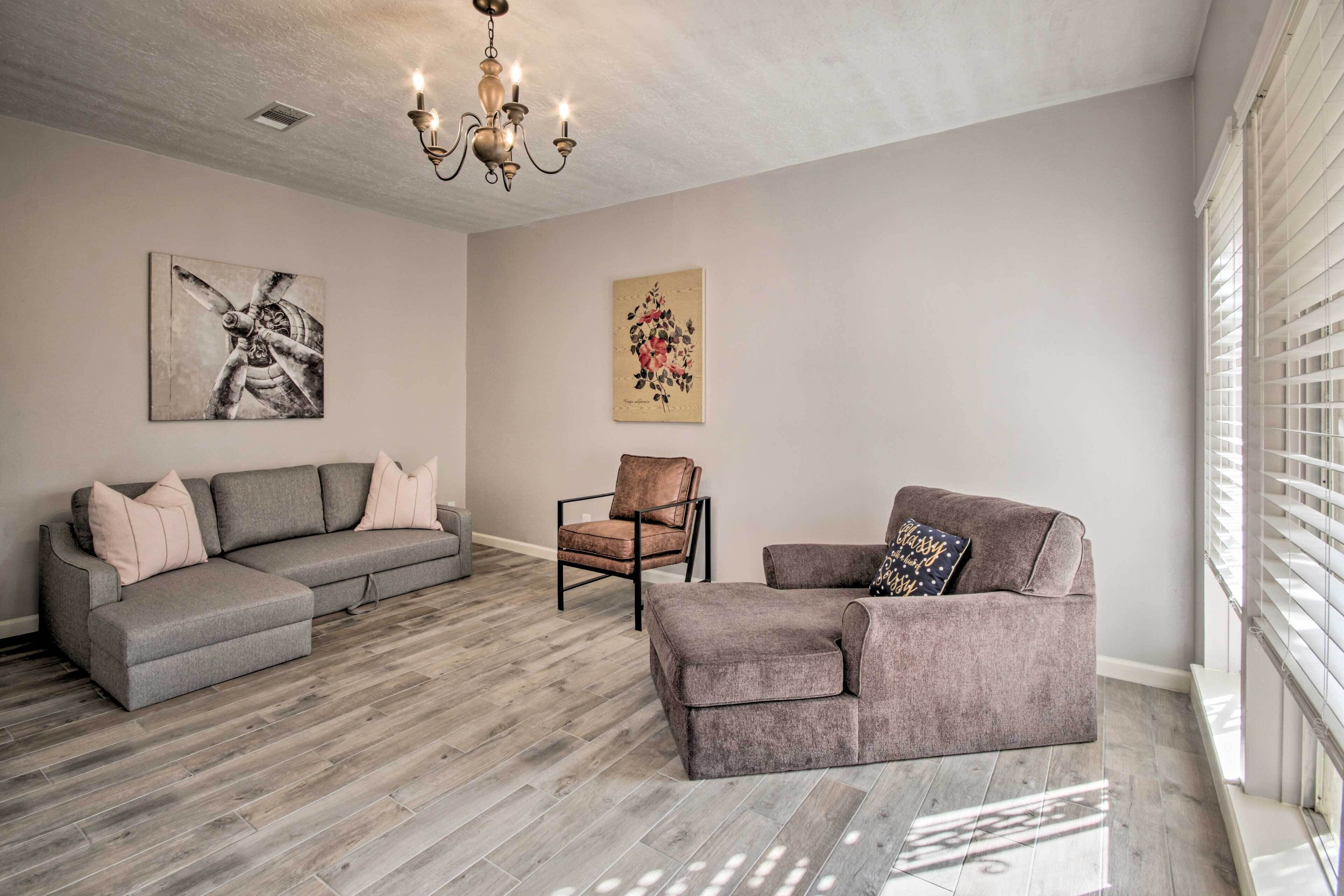 A living room with a grey sectional sofa, a brown armchair, and a small sofa with a decorative pillow. There is a chandelier on the ceiling and artwork on the walls.