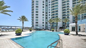 A swimming pool with a curved edge, surrounded by palm trees and lounge chairs, with a high-rise building in the background. - Luxurious Daytona Beach Condo w/ Resort Amenities! (Holly Hill)