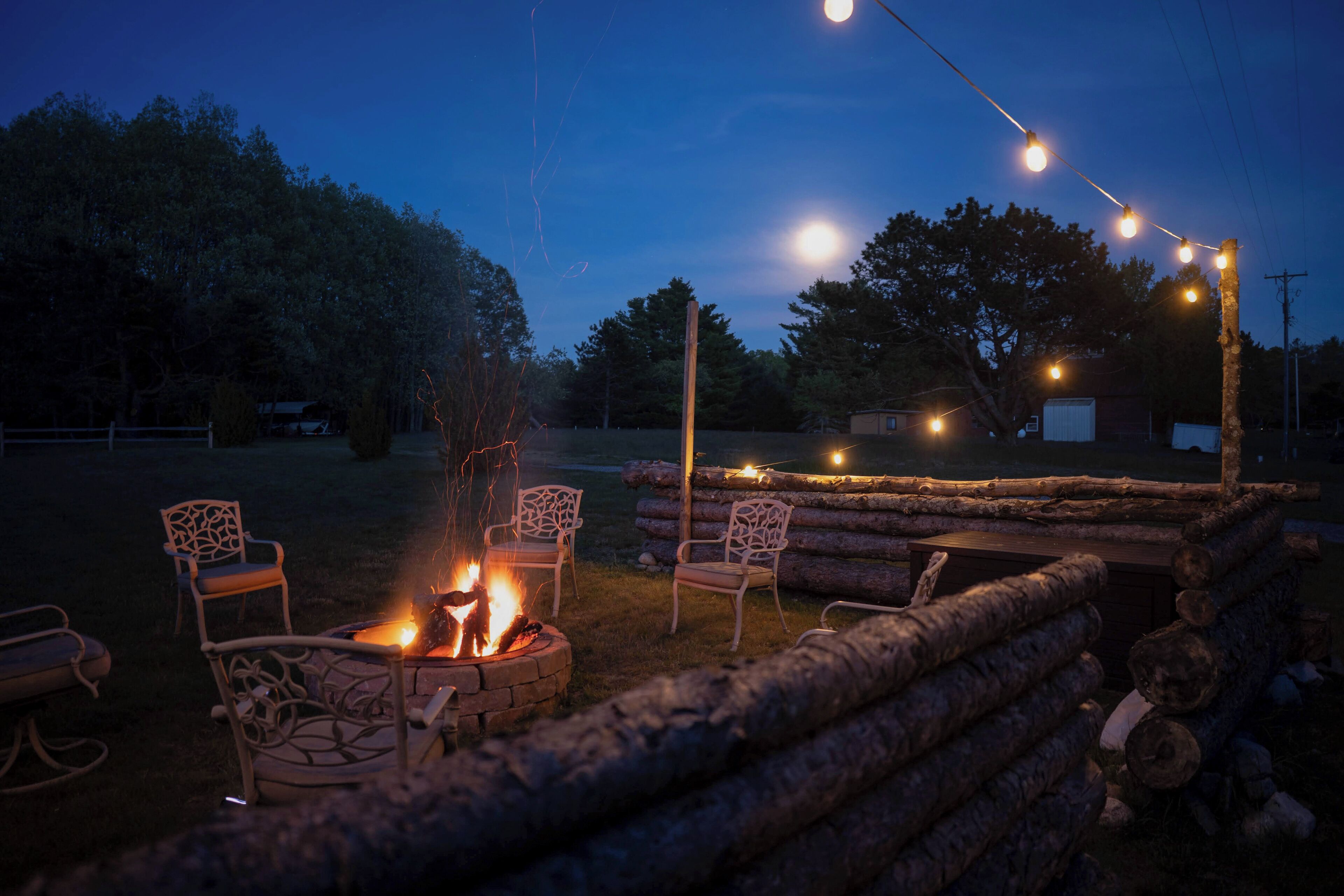 A fire pit area with chairs and logs, surrounded by trees and a clear sky.