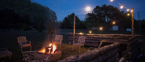 A fire pit area with chairs and logs, surrounded by trees and a clear sky.