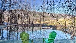 Two green Adirondack chairs facing a calm lake with a wooden dock and trees in the background.
