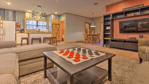 A living room with a checkered game table, sofas, a TV, and a kitchen area with a window.
