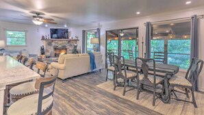 A spacious living room with a fireplace, a dining area with a table and chairs, and large windows offering a view of the outdoors. - Beautiful Connestee Falls Home w/ Porch in Brevard (Brevard)