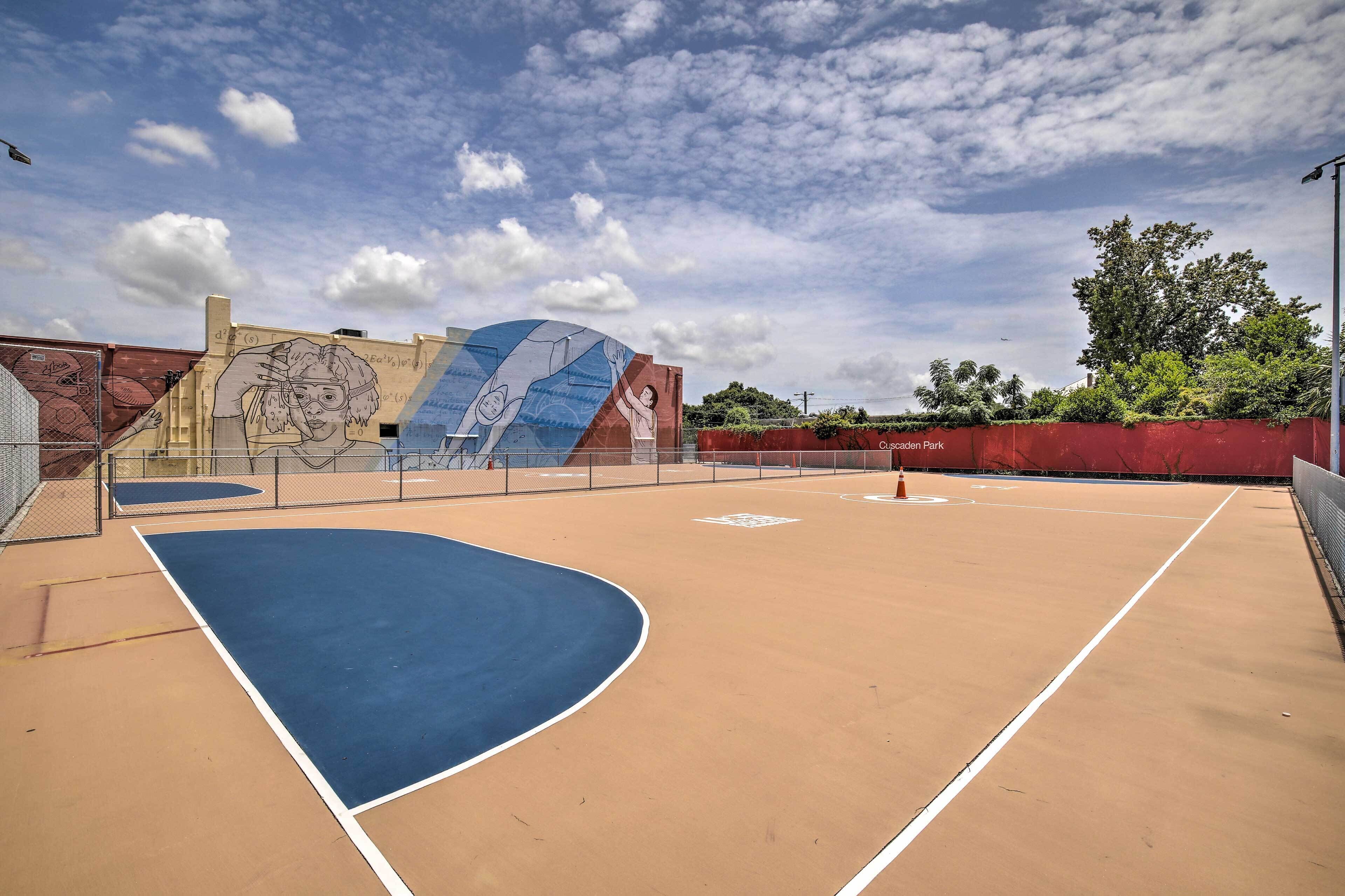 An outdoor basketball court with a mural on the wall, surrounded by a fence and trees.