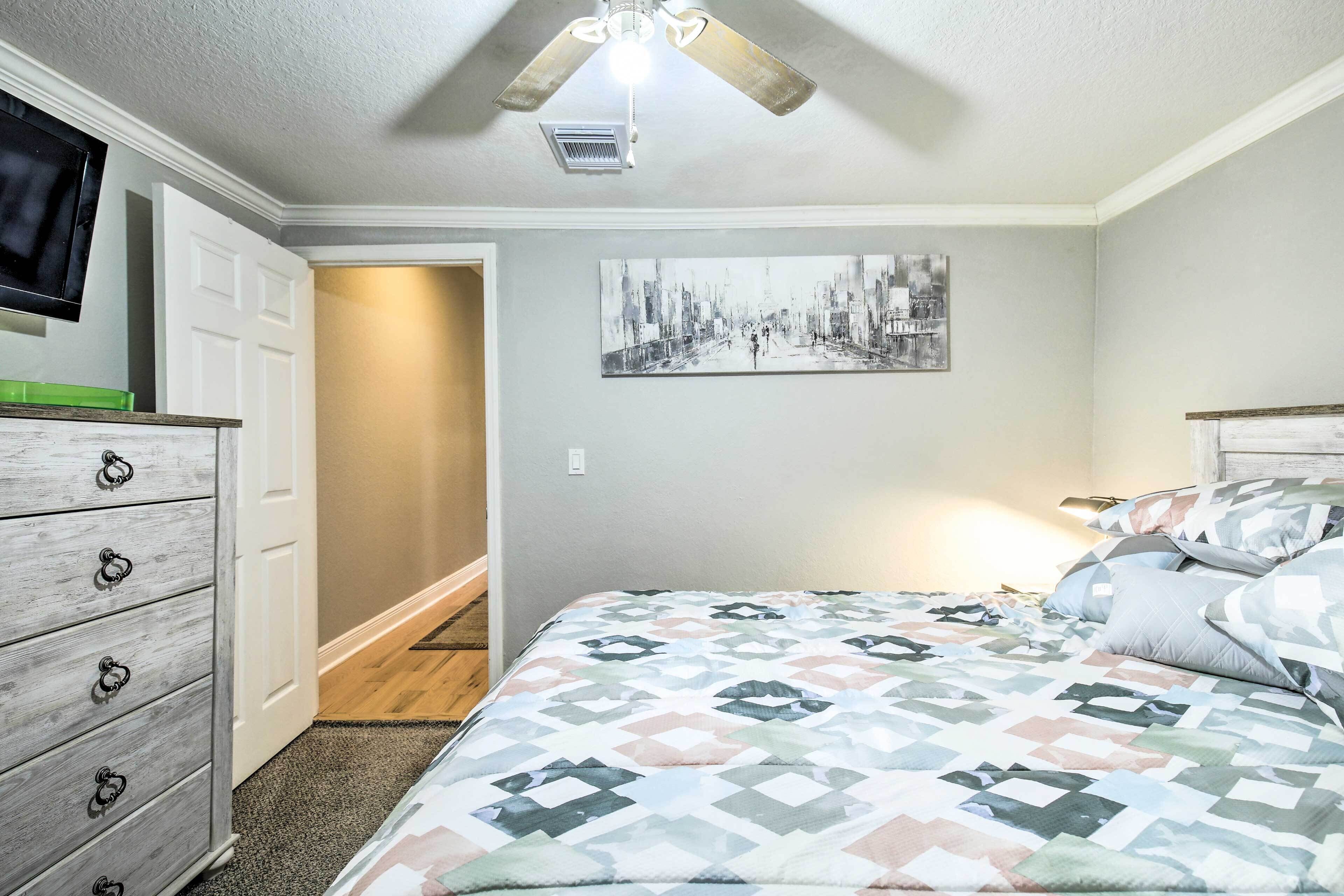 A bedroom with a bed, a dresser, a ceiling fan, a painting, and a television.