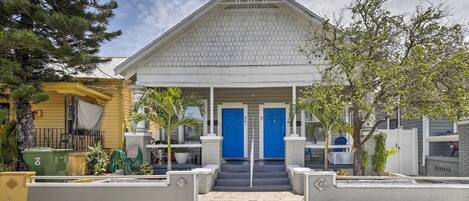 A two-story building with a white exterior and blue doors, featuring a front porch and a small garden area.