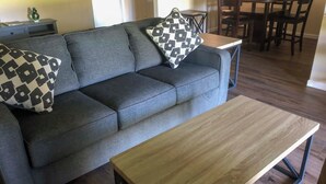 Un canapé gris agrémenté de deux coussins à motifs, une table basse en bois et un coin repas avec des chaises et une table en arrière-plan.