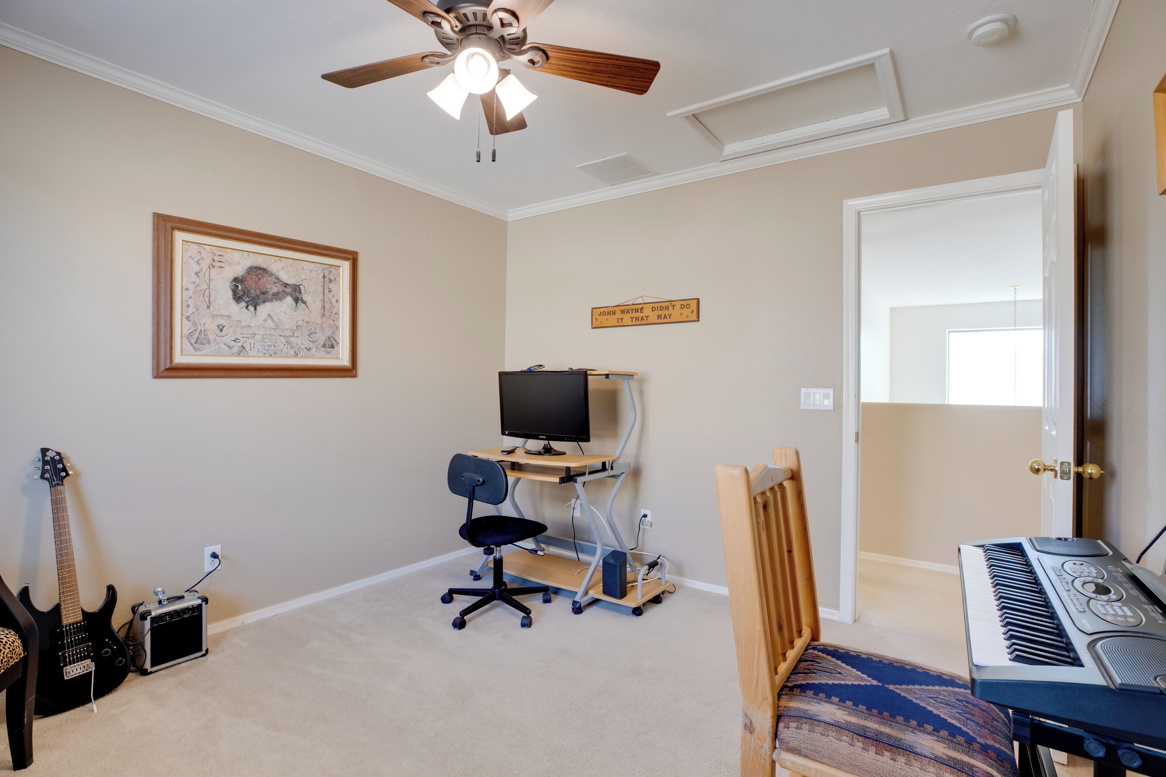 Une pièce avec un ventilateur de plafond, un tableau encadré, un bureau avec un ordinateur et une chaise, un clavier, une guitare et un petit amplificateur.