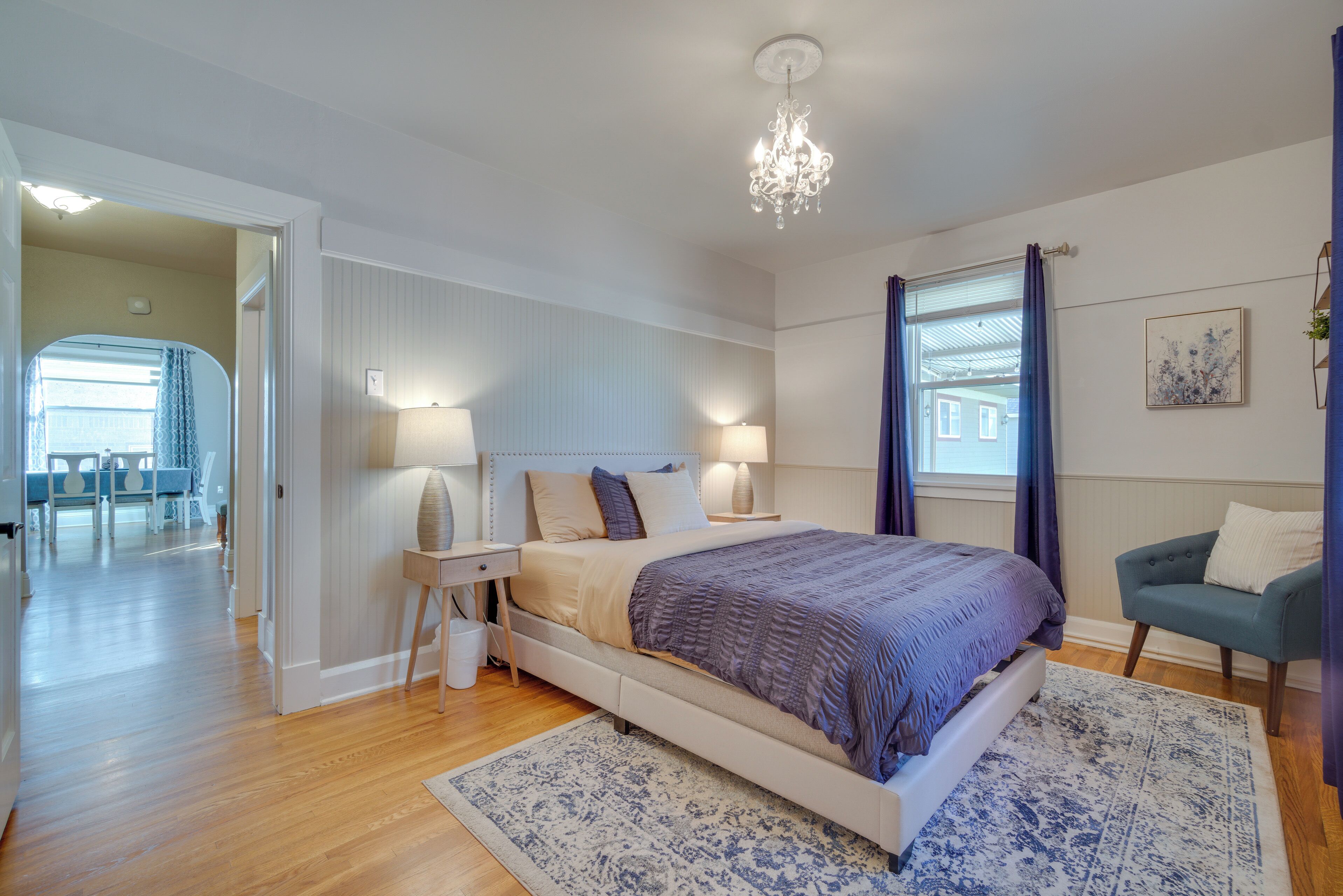 A bedroom with a large bed, bedside lamps, a chandelier, a blue chair, and a view of the outside.