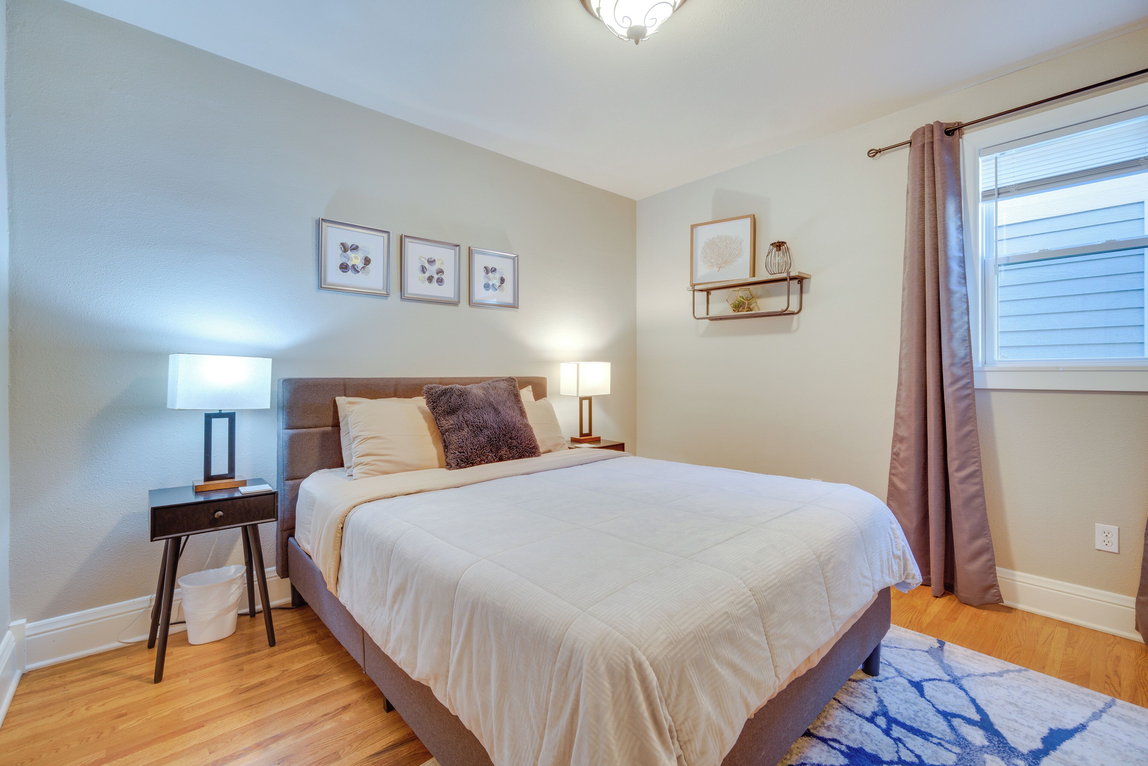 Un dormitorio con una cama grande, dos mesitas de noche con lámparas, un cubo de basura blanco pequeño, un estante de pared con adornos y una ventana con cortinas.