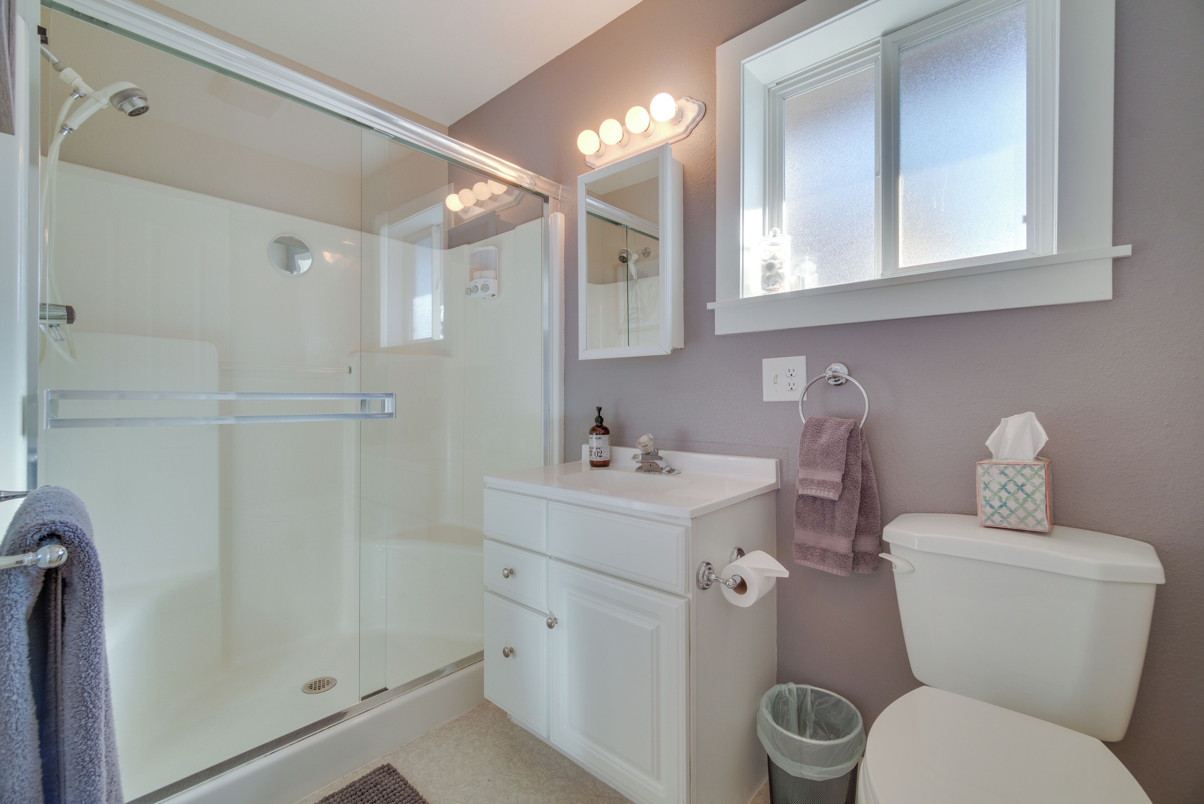 A bathroom with a glass shower enclosure, a vanity with a mirror, a toilet, and a window with lights above it.