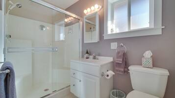A bathroom with a glass shower enclosure, a vanity with a mirror, a toilet, and a window with lights above it.
