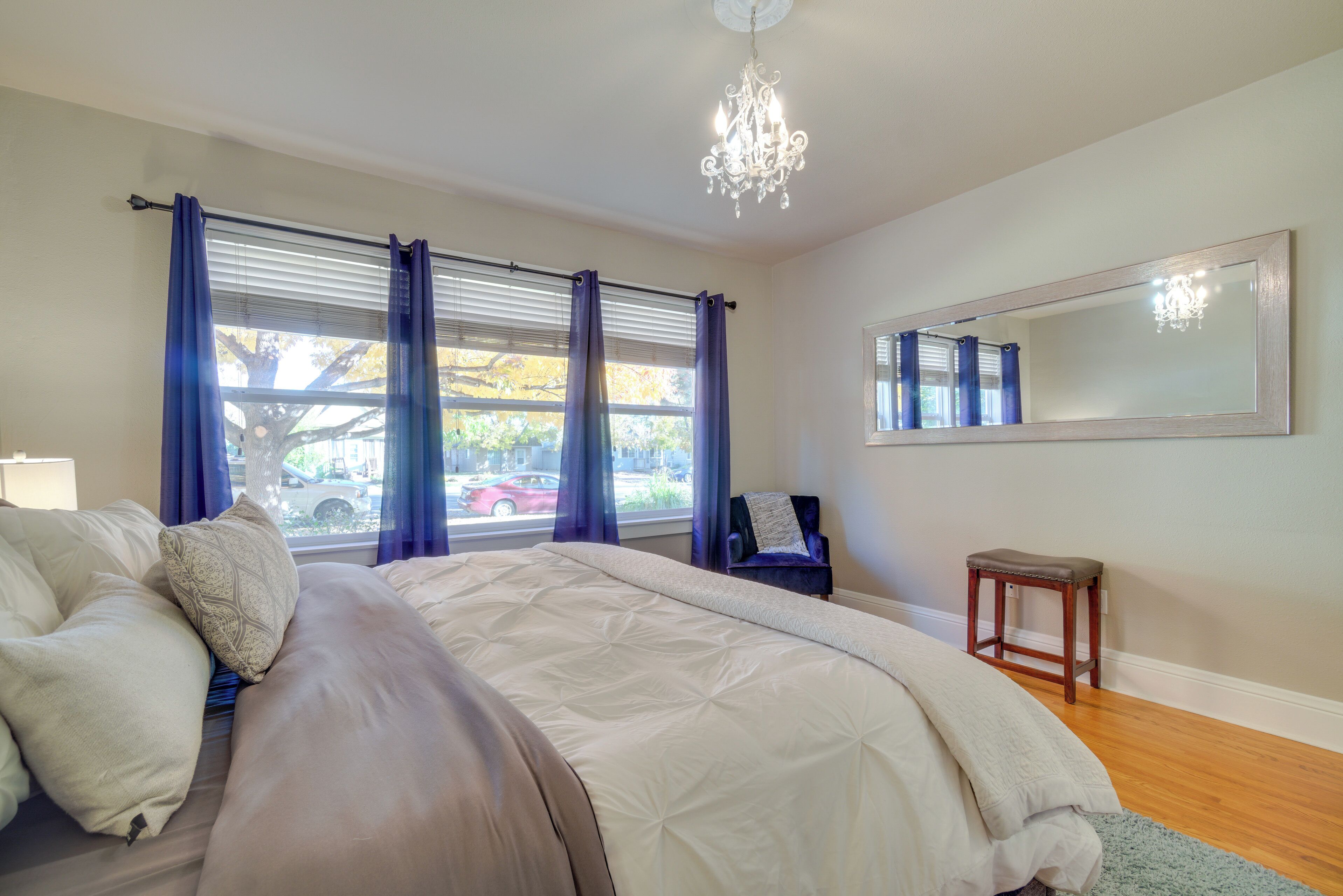Ein Schlafzimmer mit einem großen Bett, einem Kronleuchter, einem Fenster mit Vorhängen, einem Sessel und einem kleinen Hocker.
