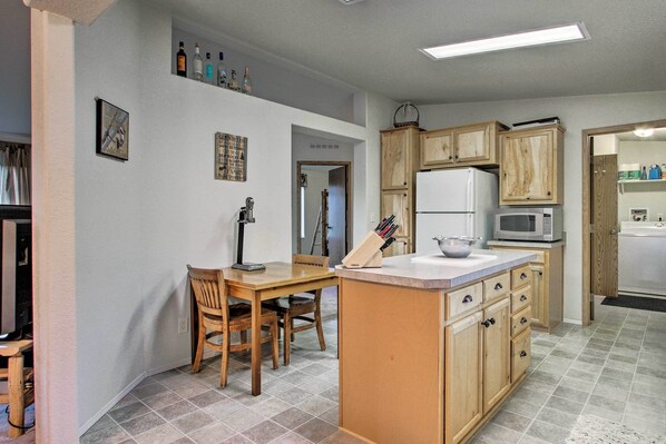 A kitchen with wooden cabinets, a refrigerator, a microwave, and a dining area with a table and chairs. - Craig Area Fly Fishing Hot Spot: Walk to River! (Cascade)