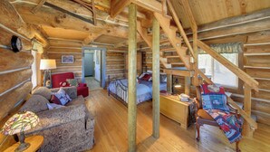 Interior de una cabaña de troncos con una escalera de madera, una sala de estar acogedora con un sofá y una lámpara, y un dormitorio con cama y ventana.