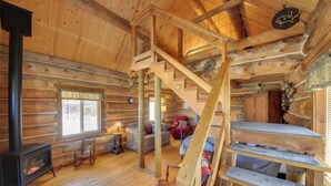 Interior de una cabaña de troncos con una escalera central, una chimenea, una zona de estar acogedora y una ventana con persianas.