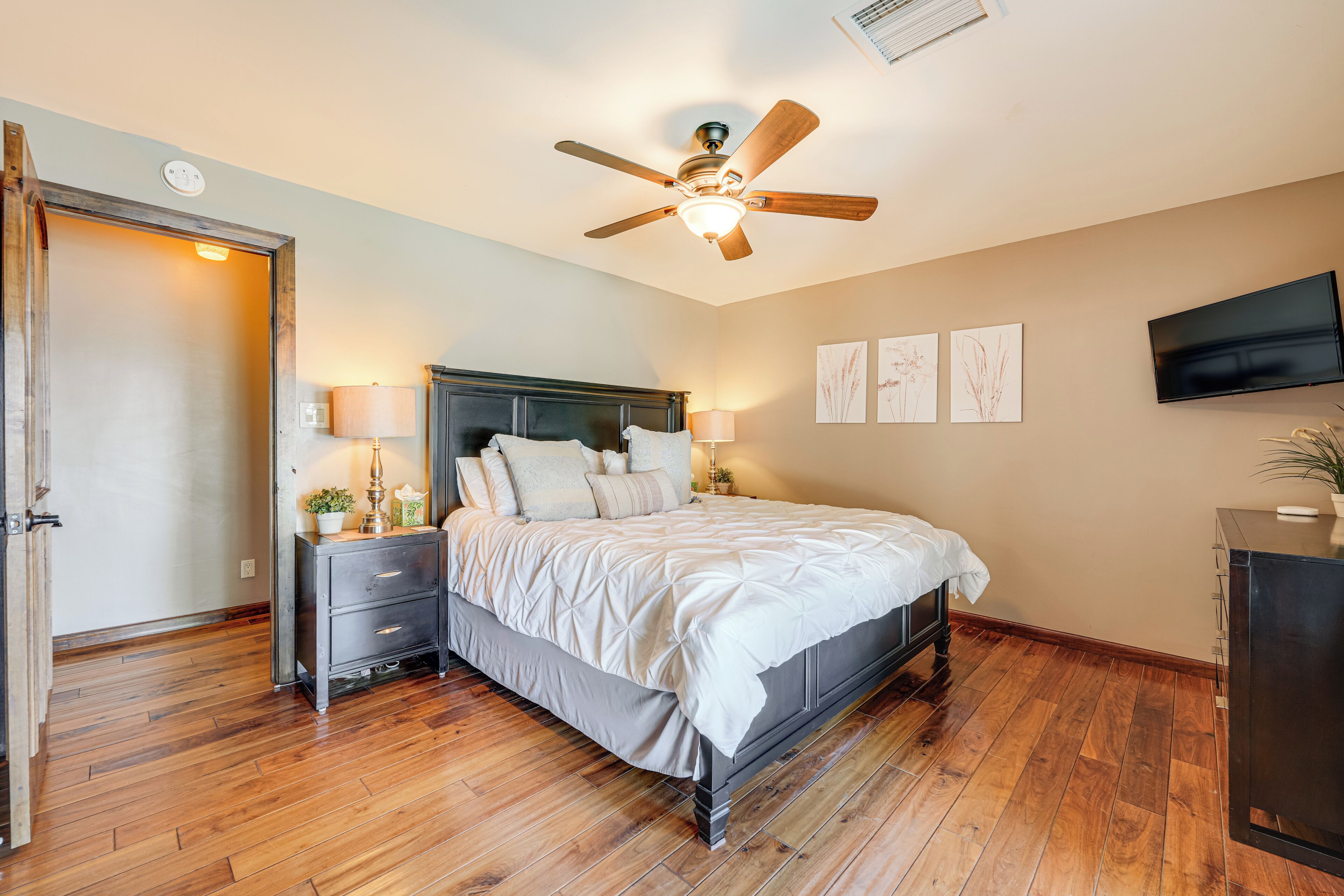 A bedroom with a wooden floor, a bed with white bedding, a ceiling fan, bedside lamps, and a TV on the wall.