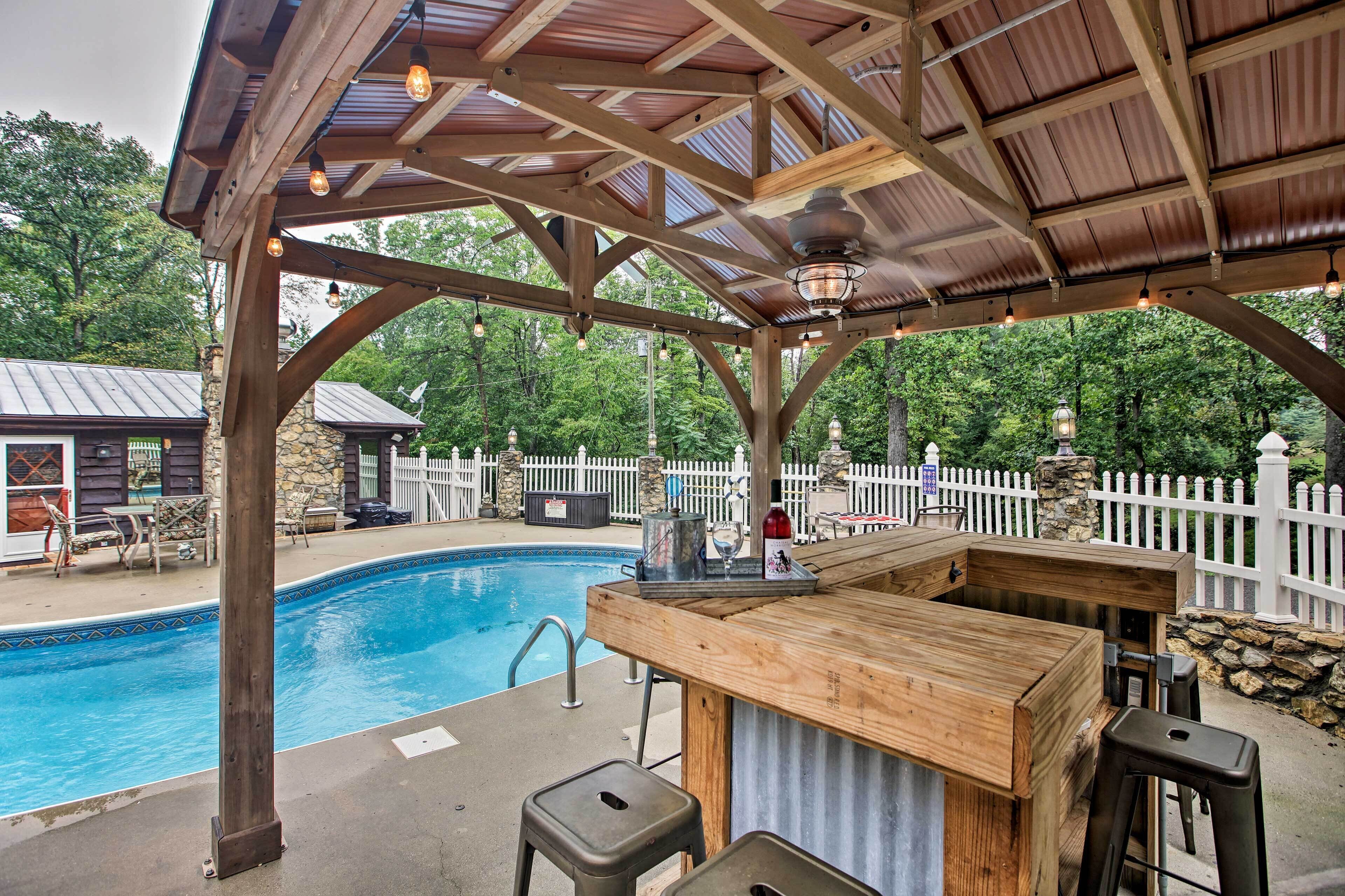 A wooden poolside bar with stools, a pool, and a white picket fence in the background.