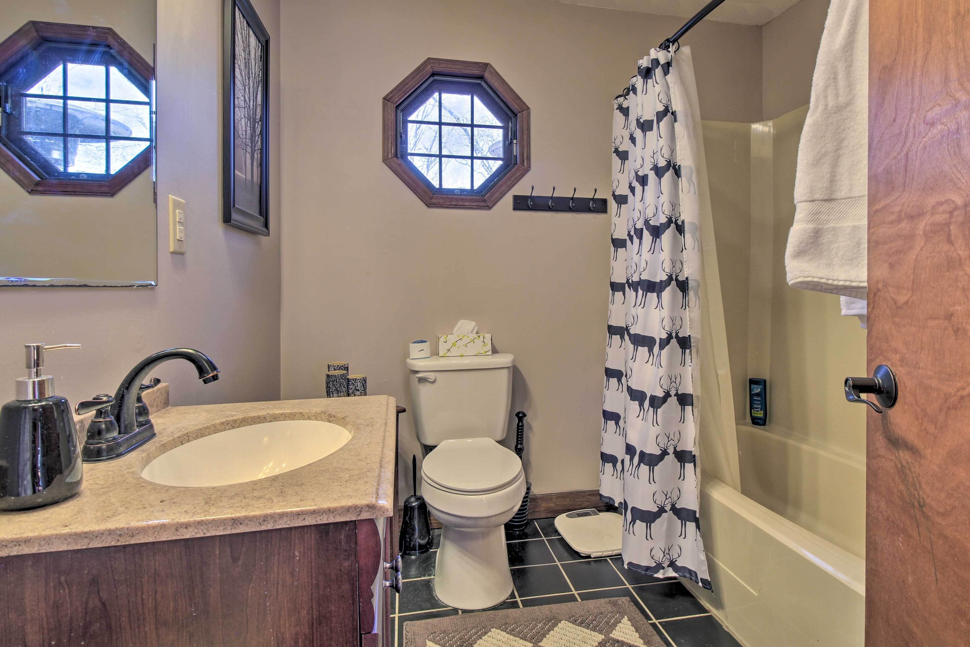 Un baño con lavamanos, inodoro y ducha. Cuenta con dos ventanas y un perchero para toallas.