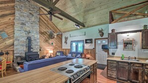 Interior rústico de una cabaña con pared de piedra, techo de madera, una isla central con estufa, un sofá y un comedor.