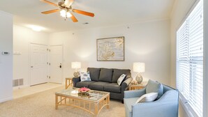 A living room with a grey sofa, a blue armchair, a glass coffee table, two side tables with lamps, a ceiling fan, and a window with blinds. - 4 Mi to Beaches: Luxe Coastal Resort Condo (Lewes)