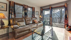 A living room with a brown leather sofa, a glass coffee table, and a view of a deck through large windows. - 1 Mile to Golfing: Porch w/ Fireplace + Mtn Views! (Maggie Valley)