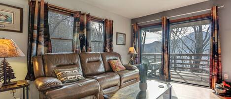A living room with a brown leather sofa, a glass coffee table, and a view of a deck through large windows.