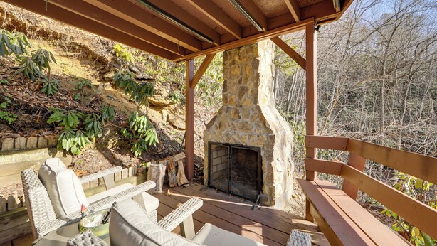 An outdoor patio with wicker furniture, a stone fireplace, and a wooden deck overlooking a wooded area.