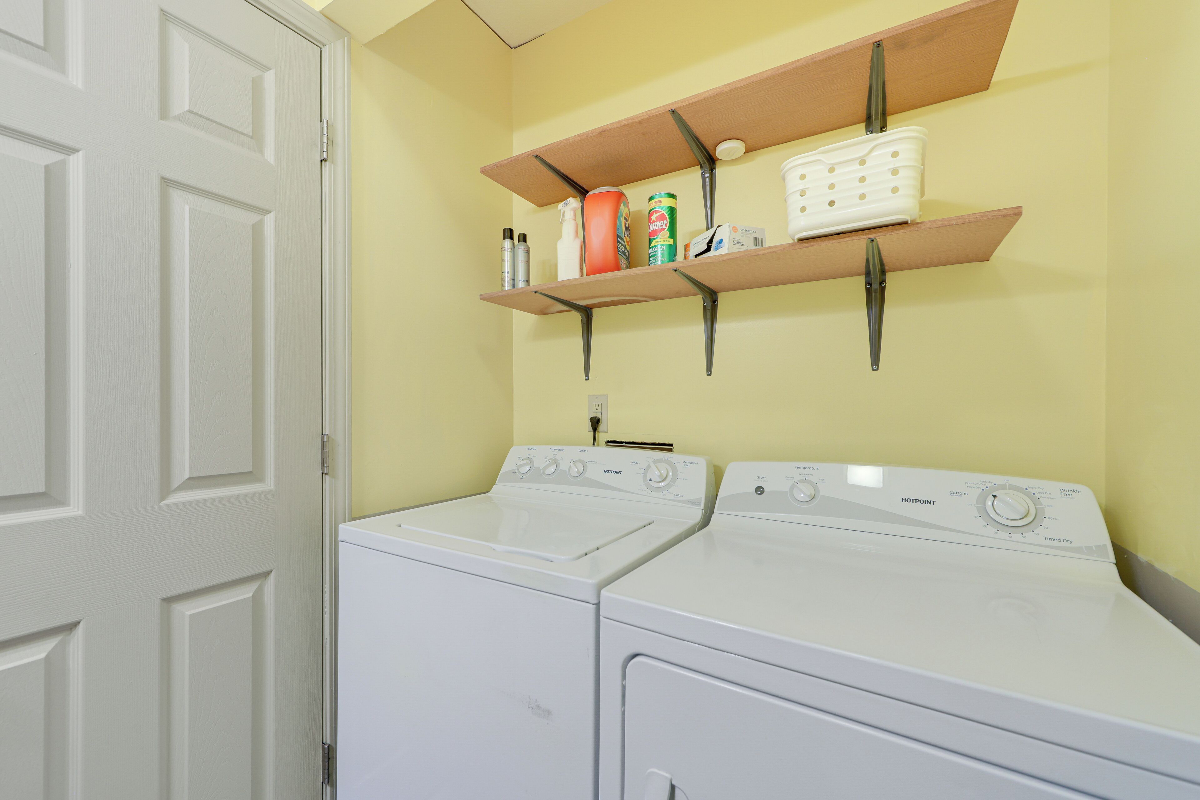 A laundry room with two white front-loading washers and dryers, a shelf above holding cleaning supplies, and a white door.