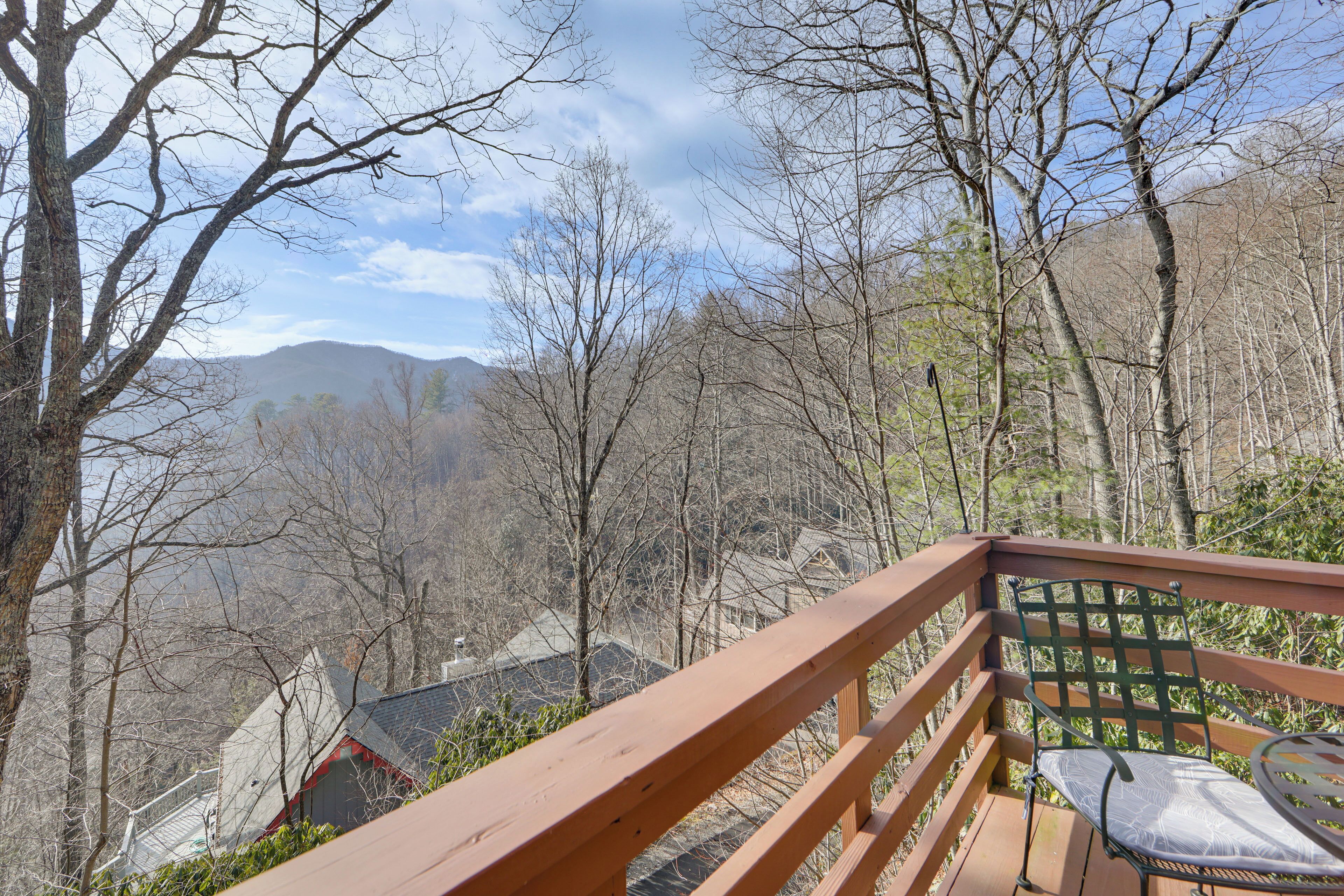 A wooden deck with a metal chair and table, overlooking a mountainous landscape with bare trees and a house in the distance.