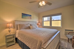 A bedroom with a large bed, two bedside lamps, a ceiling fan, a window with a view, and a chair. - Walk to Ocean: Cozy Condo in North Myrtle Beach! (North Myrtle Beach)