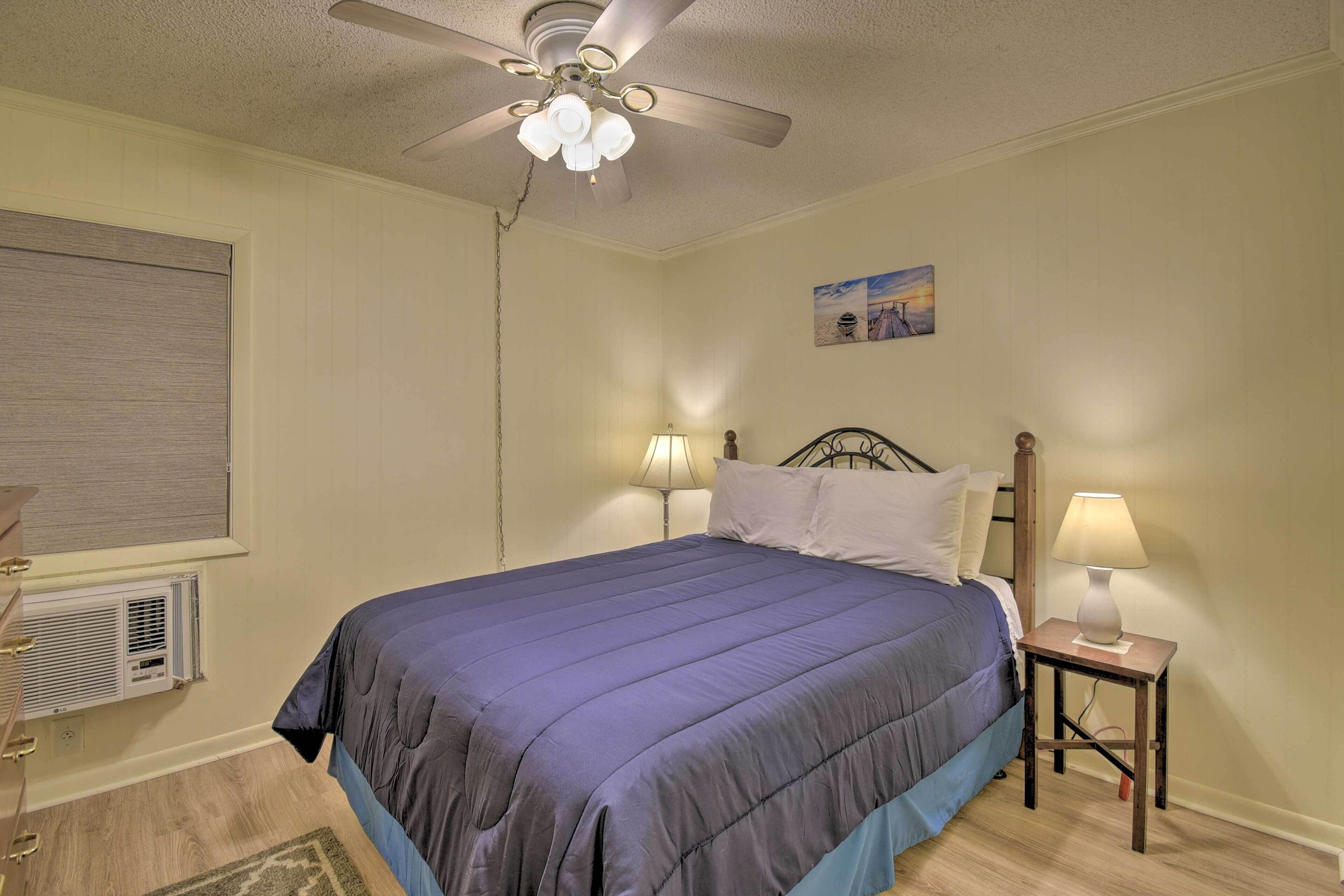 A bedroom with a bed, ceiling fan, wall-mounted air conditioner, bedside lamps, and a painting on the wall.