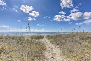 A sandy path leading to a beach with tall grasses and a clear blue sky. - Walk to Ocean: Cozy Condo in North Myrtle Beach! (North Myrtle Beach)