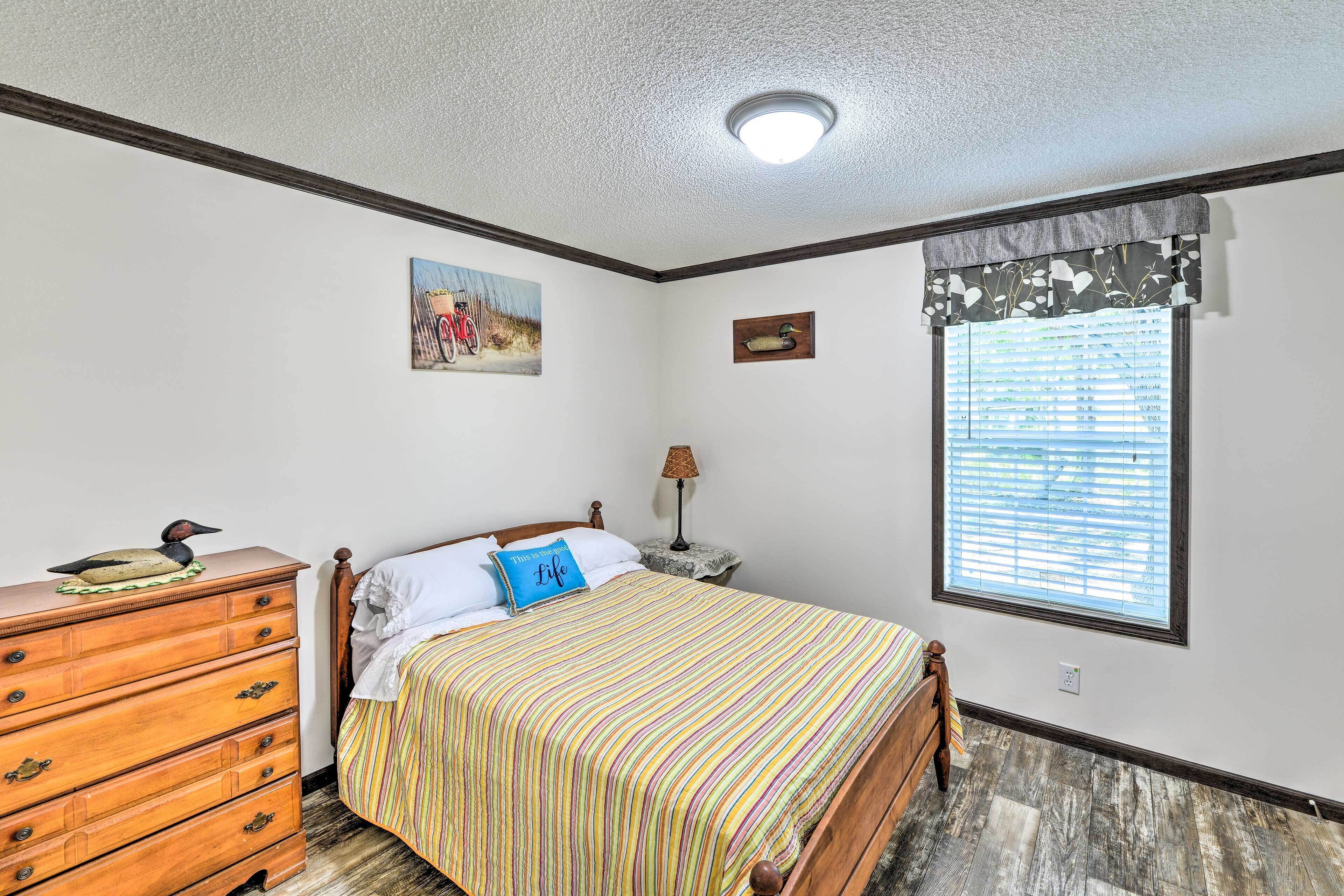 A bedroom with a wooden dresser, a bed with a striped bedspread, a window with blinds, and a painting on the wall.