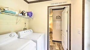 A laundry area with two white washers and dryers, a shelf with laundry supplies, and a hallway leading to a room with a wooden floor. - 'pelican Place' Coastal Cottage Walk to Back Sound (Harkers Island)