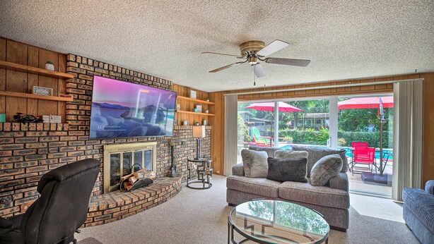 A living room with a brick fireplace, a flat-screen TV mounted on the wall, a ceiling fan, and a view of a pool and outdoor furniture through large sliding glass doors. - Spacious Family Glenwood Estates Home w/ Hot Tub! (Glenwood)