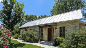 A stone house with a metal roof, a covered porch, and a well-maintained garden with pink flowers. - Stonebridge 2: 2 Blocks to Main, Shared Hot Tub (Fredericksburg)