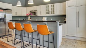 A modern kitchen with white cabinets, a stainless steel refrigerator, a microwave, and a bar area with orange stools. - Pet-friendly Snowbird Haven w/ Pool in Gold Canyon (Gold Canyon)