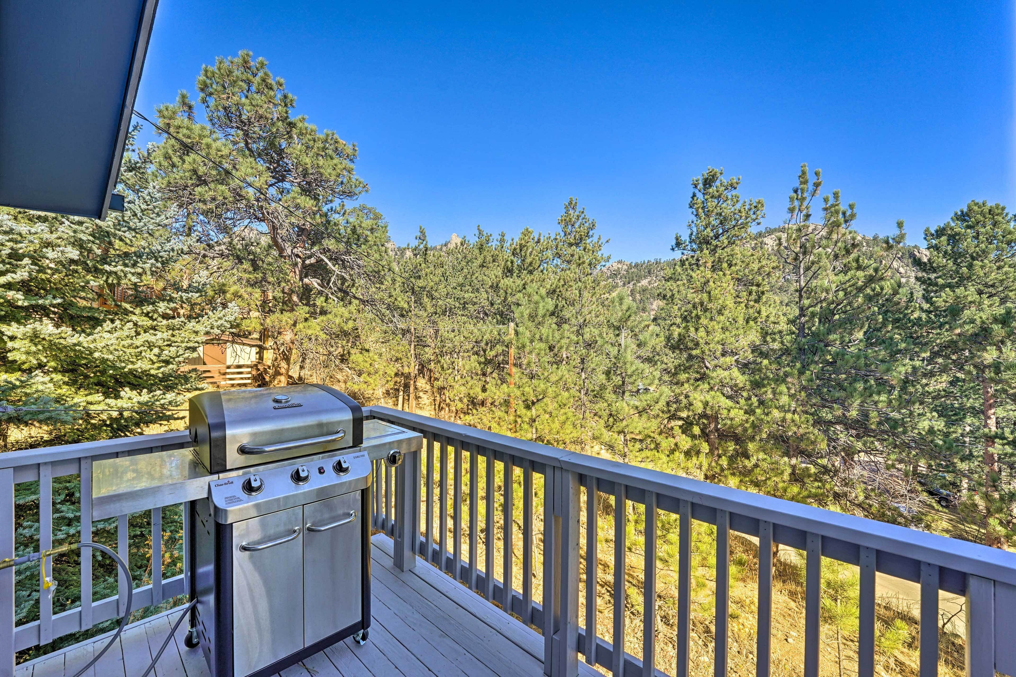 A deck with a stainless steel grill, surrounded by a wooden railing, overlooking a forested area.