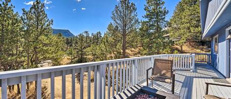 A wooden deck with a fire pit, white railings, and a view of pine trees and a mountain.