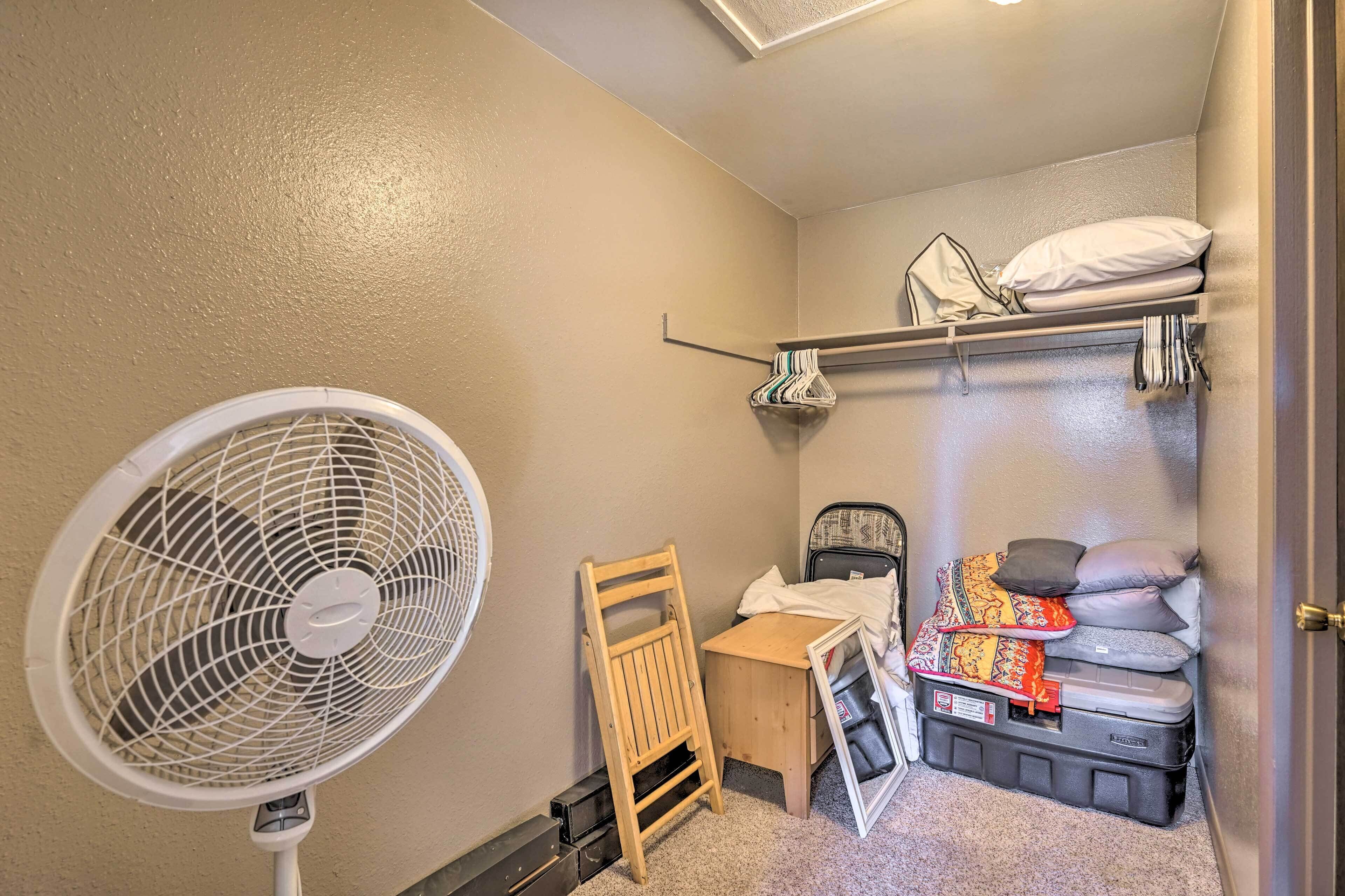 A compact room with a standing fan, a wooden chair, a small desk, and a bed with storage.