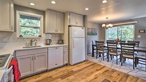 A modern kitchen with white cabinets, a built-in dishwasher, and a refrigerator. There is a dining area with a wooden table and chairs, and a window providing a view of trees. - Luxe Updated Home w/ Grill + Hot Tub: 4 Mi to Rmnp (Estes Park)