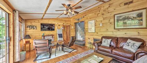 A cozy cabin interior with a wooden ceiling, a fireplace, a leather sofa, a dining table, and a staircase leading to an upper level.
