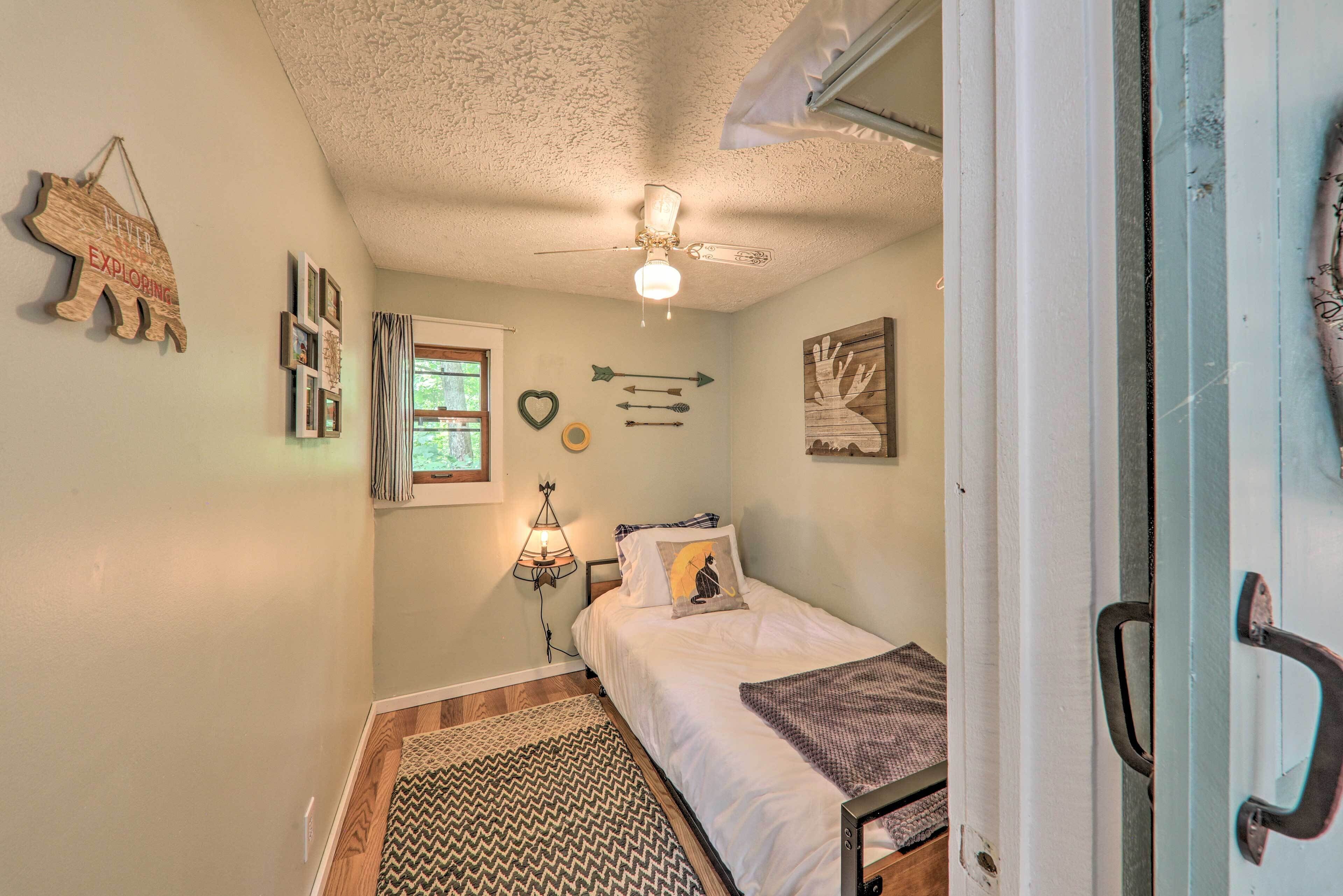 A small bedroom with a bed, a ceiling fan, a window, and a wall-mounted shelf.