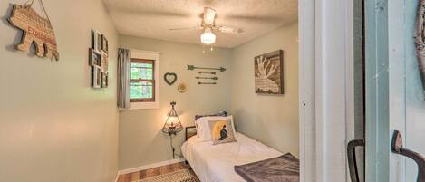 A small bedroom with a bed, a ceiling fan, a window, and a wall-mounted shelf.