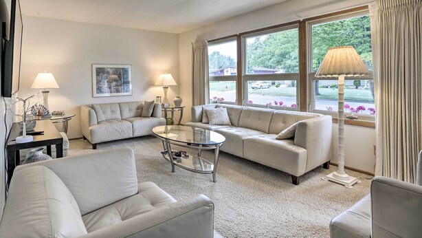 A living room with a beige sofa, a glass coffee table, and a floor lamp. There is a painting on the wall and a window with curtains.
