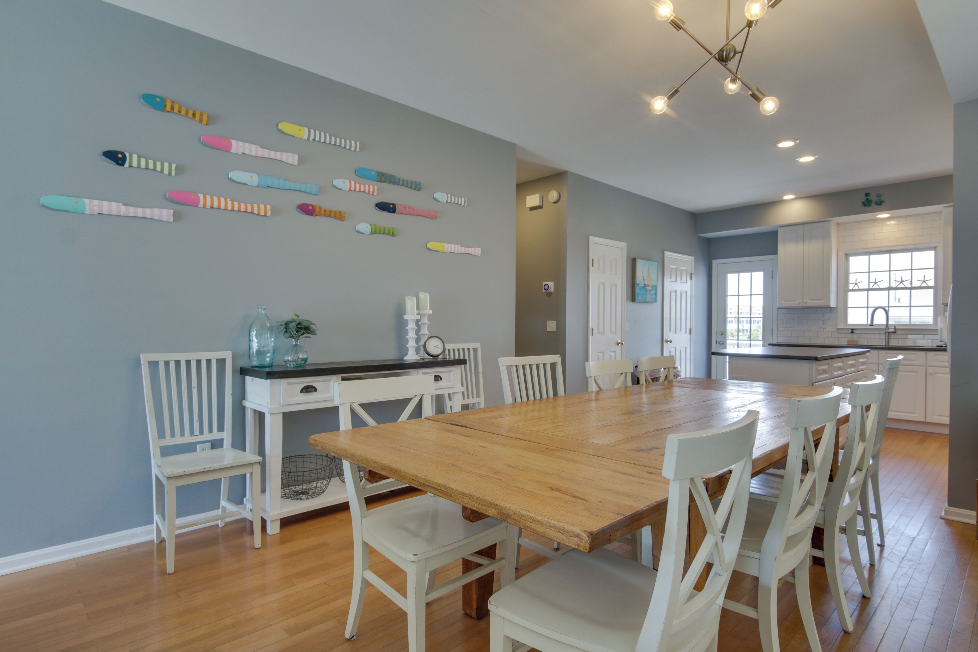 A dining area with a wooden table and white chairs, a kitchen in the background, and a wall with colorful fish decorations.